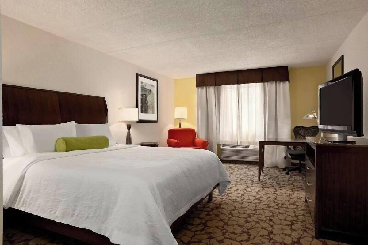 A hotel room with a large bed, a desk with a television, a chair, a lamp, and a window with curtains.