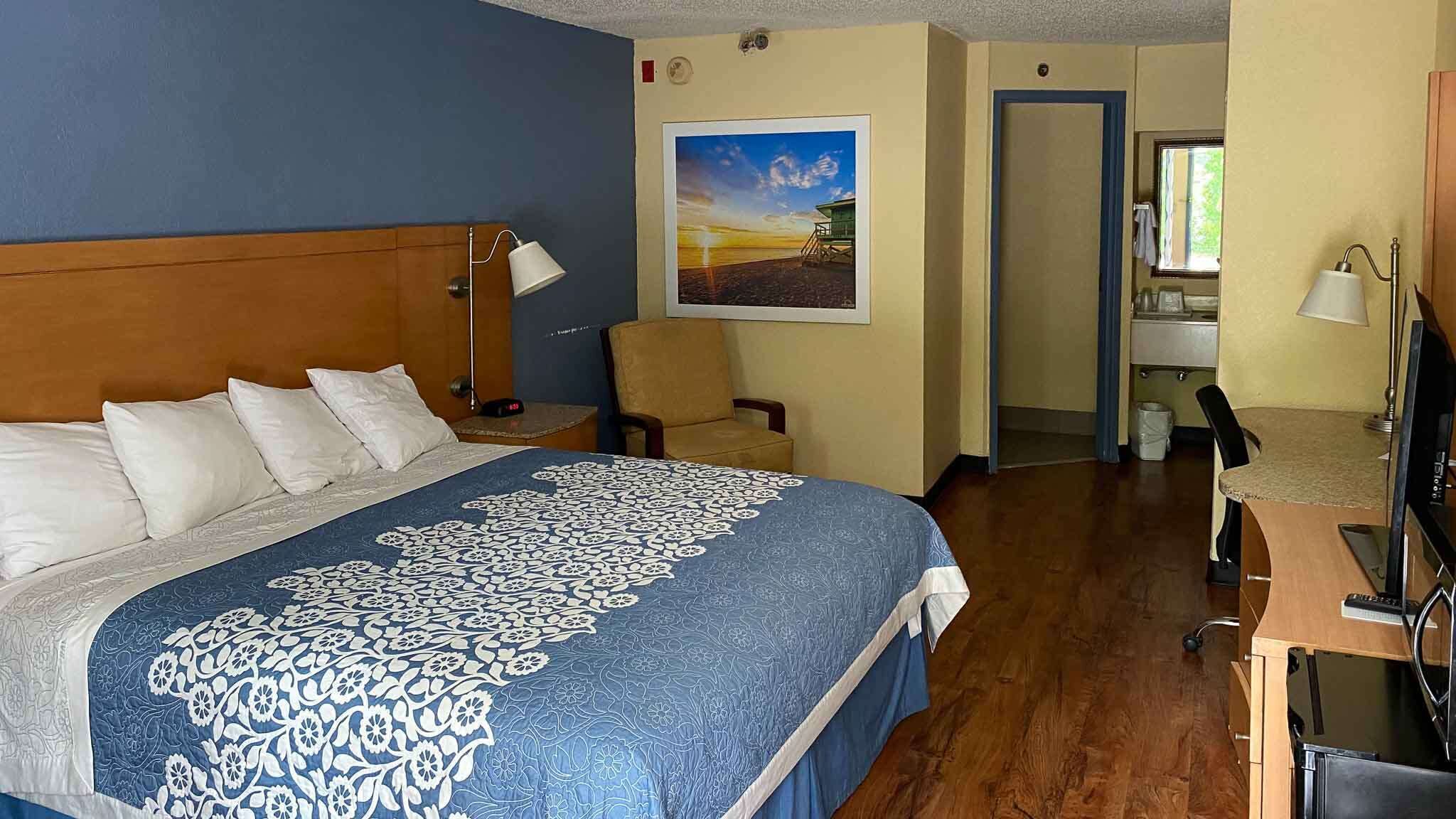 A hotel room with a large bed, a desk with a computer, a chair, and a painting on the wall.