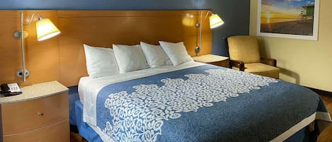 Una habitación de hotel con cama, dos lámparas de noche, una silla, un escritorio con teléfono y un cuadro en la pared.