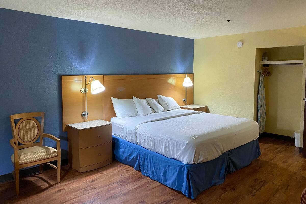 A hotel room with a wooden headboard, a bed with white linens, a nightstand with a lamp, a wooden chair, and a wooden floor.