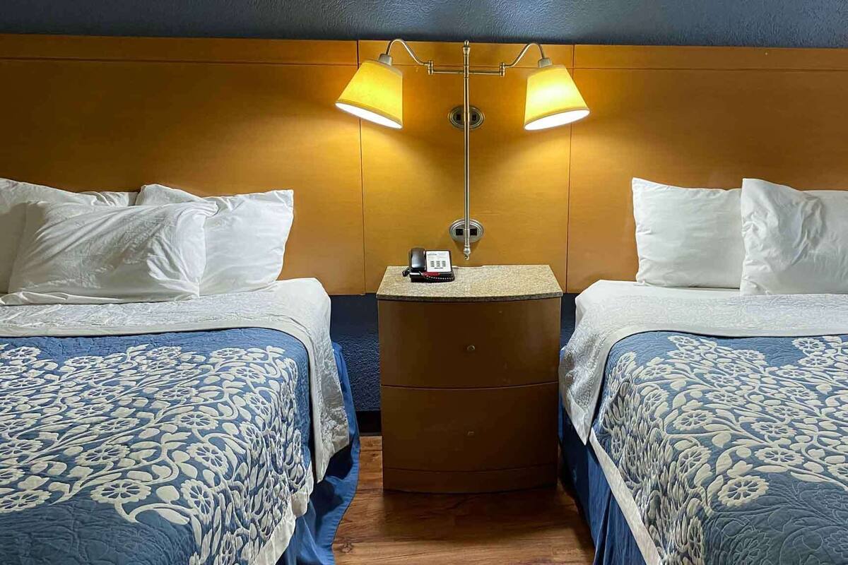 A hotel room with two beds, each with a blue floral bedspread and white pillows. There is a wooden headboard, a small wooden side table with a phone, and two wall-mounted lamps.