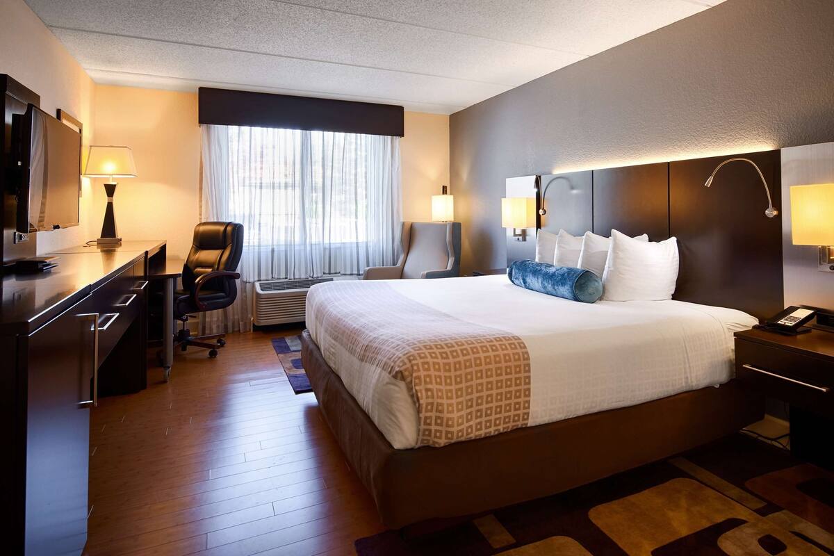 A hotel room with a large bed, a desk with a chair, a TV, and a bedside table with a lamp.
