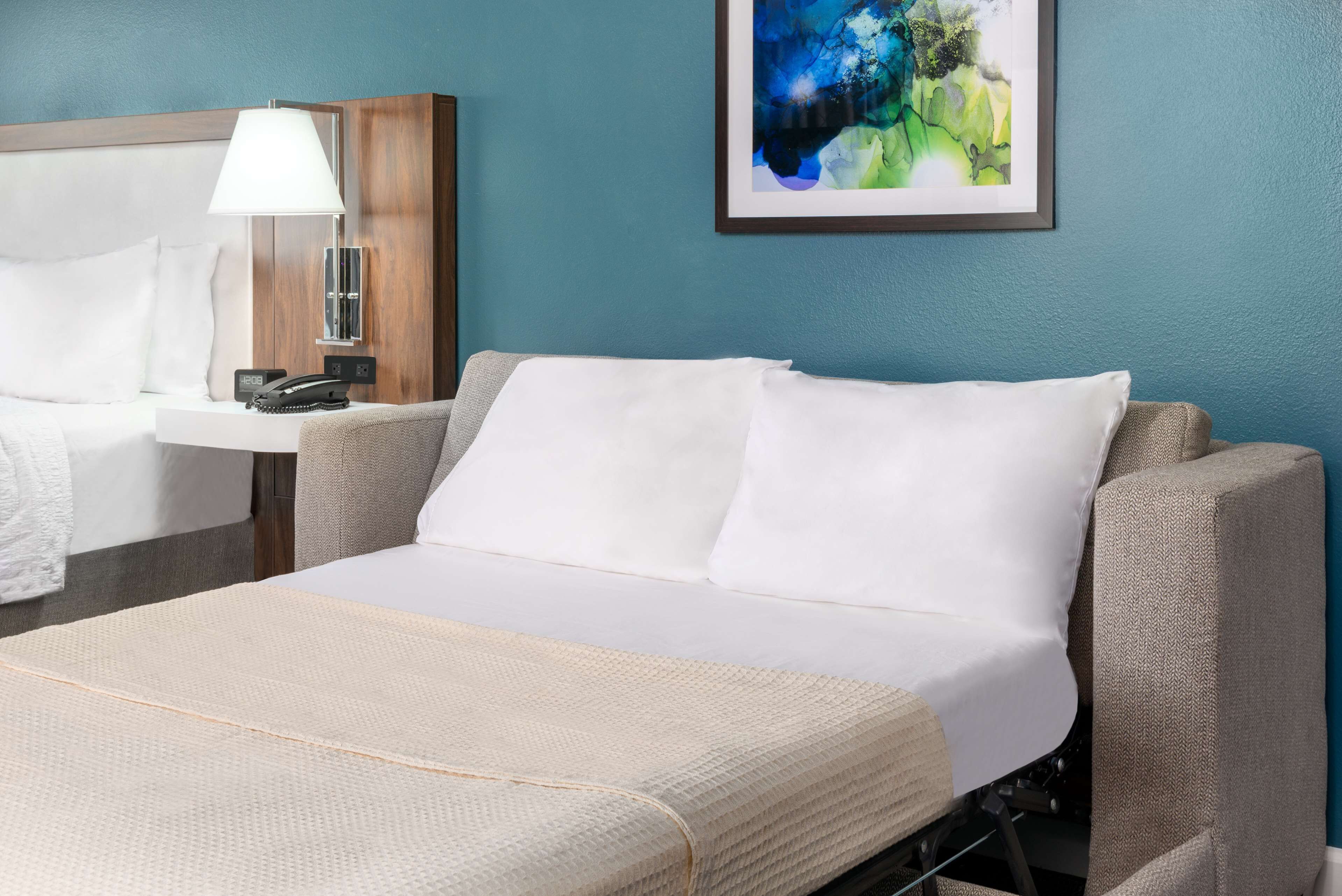 Une chambre d’hôtel avec un lit, une table de chevet avec une lampe, un téléphone et un tableau abstrait encadré accroché au mur.
