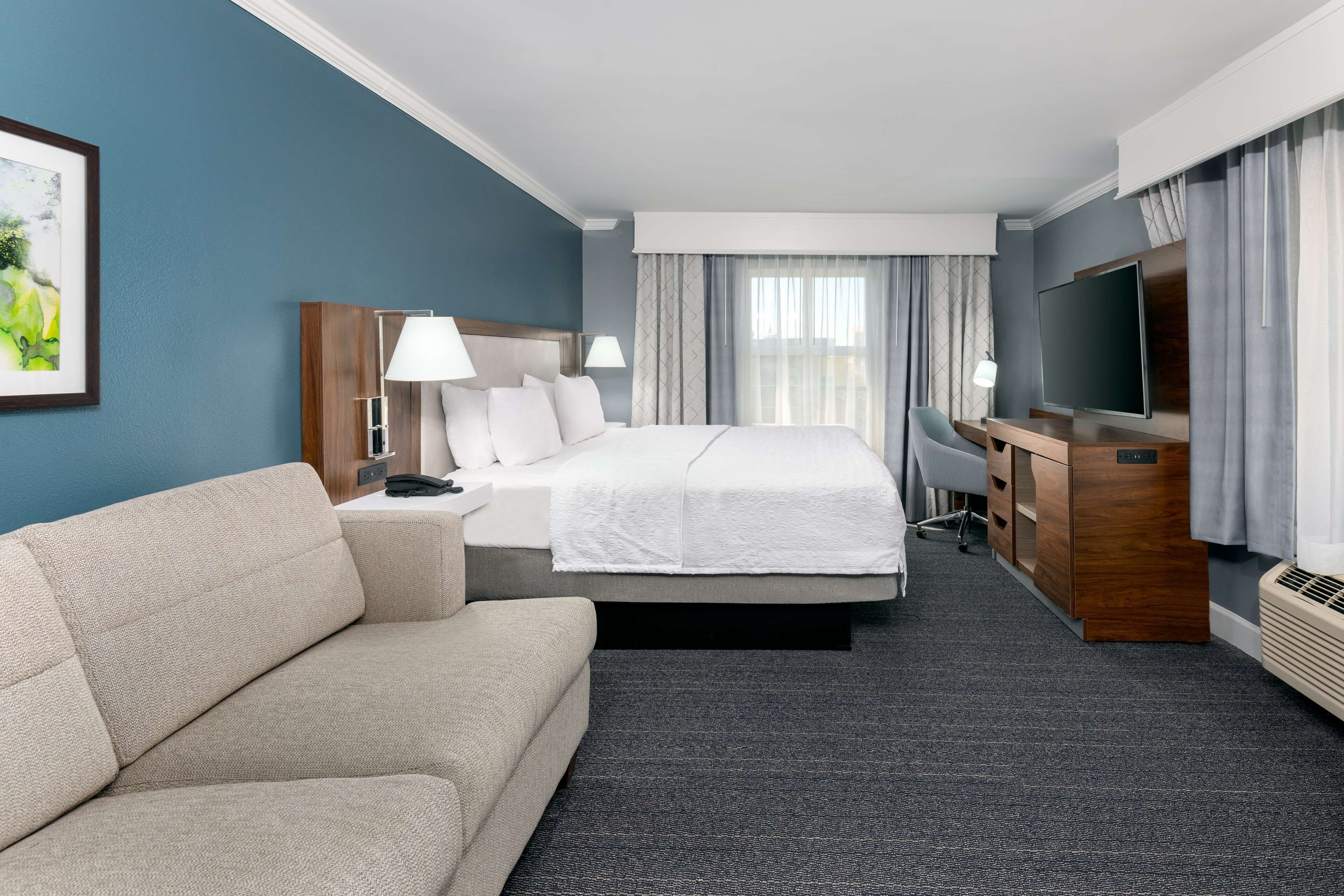 A hotel room with a bed, a sofa, a desk, a TV, and a large window with curtains.
