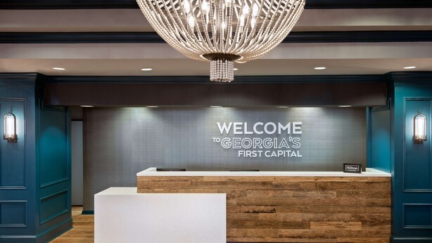 A reception desk with a "WELCOME TO GEORGIA'S FIRST CAPITAL" sign, a modern chandelier, and a wooden reception desk. - Hampton Inn Savannah-Historic District (Savannah)