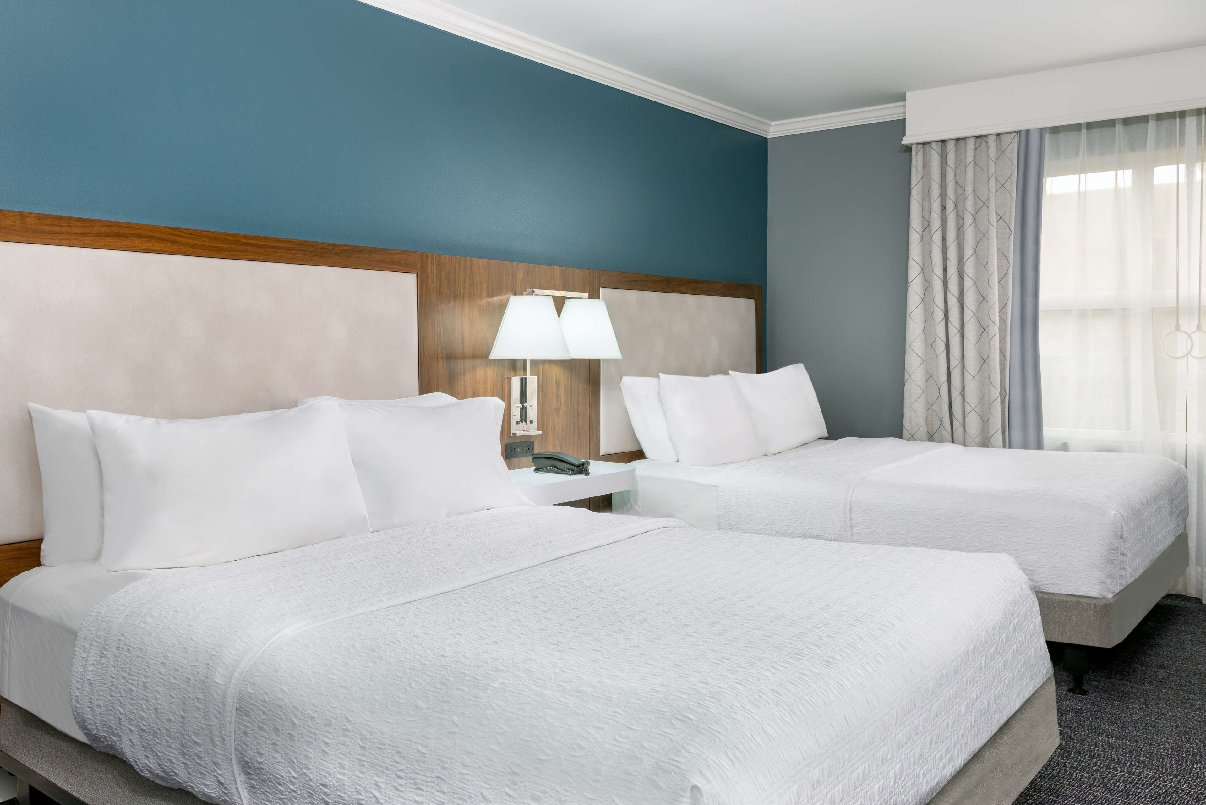 A hotel room with two beds, a nightstand with a lamp, and a window with curtains.
