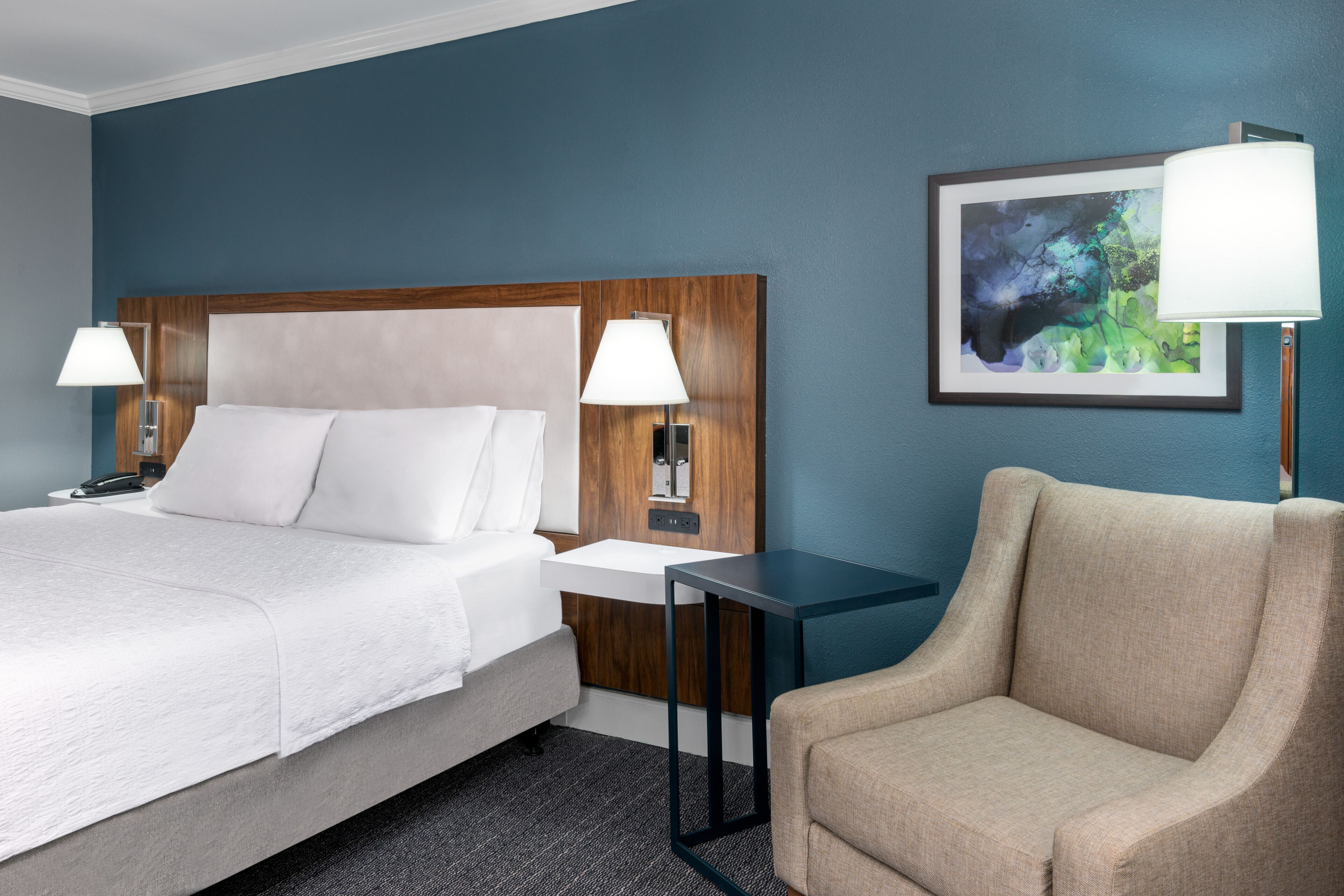 Un cuarto de hotel con una cama grande, mesitas de noche con lámparas, una silla y una obra de arte enmarcada en la pared.