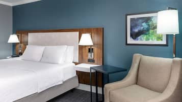 Un cuarto de hotel con una cama grande, mesitas de noche con lámparas, una silla y una obra de arte enmarcada en la pared.