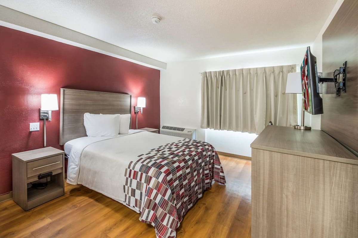 Una habitación de hotel con cama, mesitas de noche, escritorio, televisor y ventana con cortinas.