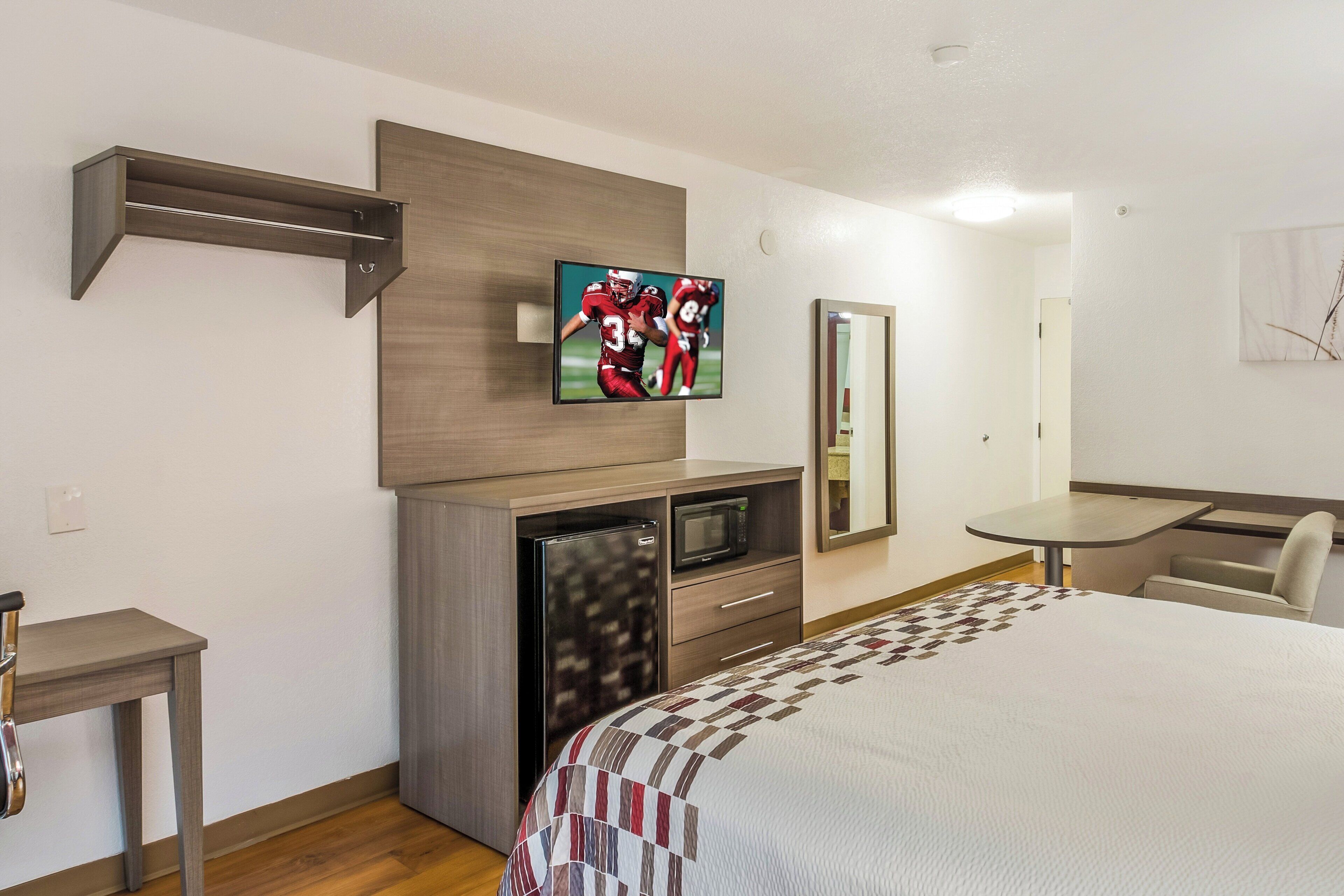 A hotel room with a bed, a television mounted on a wooden panel, a microwave, a small table, and a chair.