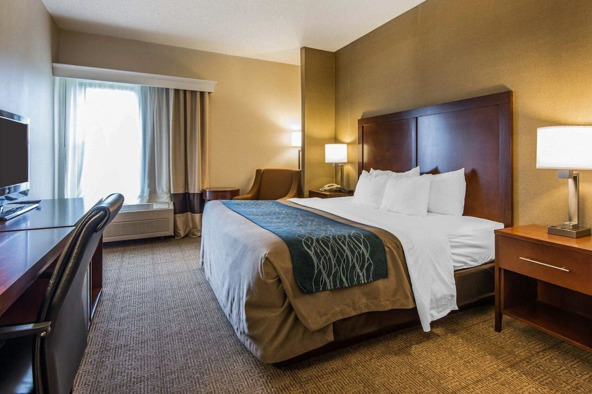 A hotel room with a bed, bedside tables, a desk with a chair, a TV, and a window with curtains.