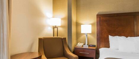 A hotel room with a bed, a chair, a nightstand with a lamp, and a small table.