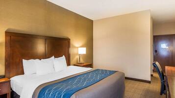A hotel room with a large bed, a nightstand with a lamp, a desk with a chair, and a door.