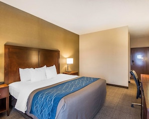A hotel room with a large bed, a nightstand with a lamp, a desk with a chair, and a door.