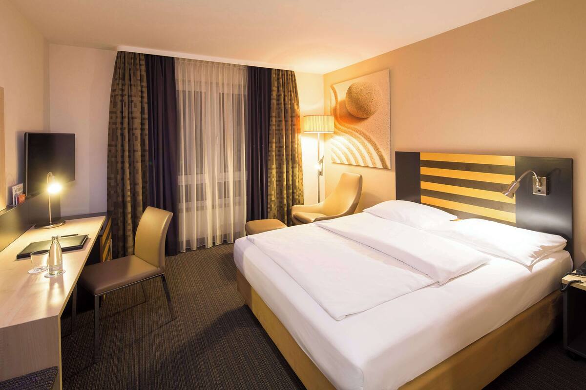 A hotel room with a bed, a desk with a lamp, a chair, and a wall-mounted phone.