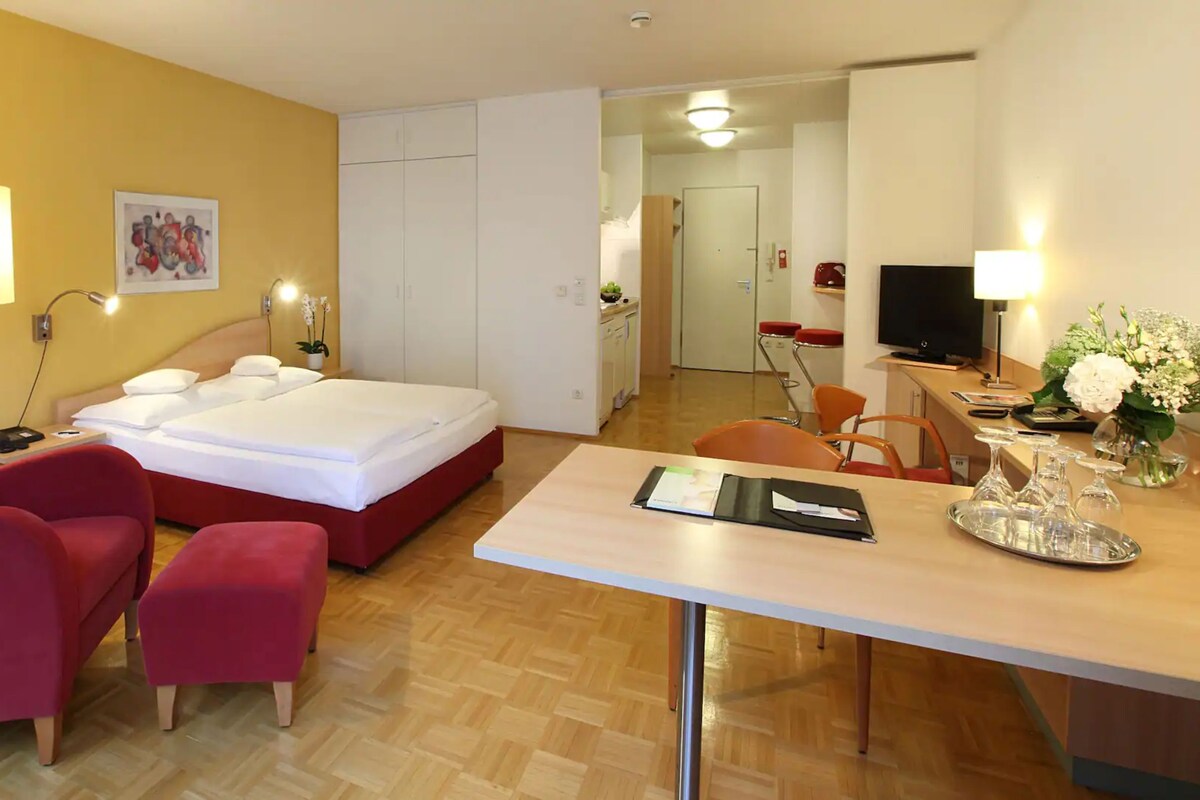 A hotel room with a bed, a desk, a chair, a small table, a lamp, a TV, and a painting on the wall.