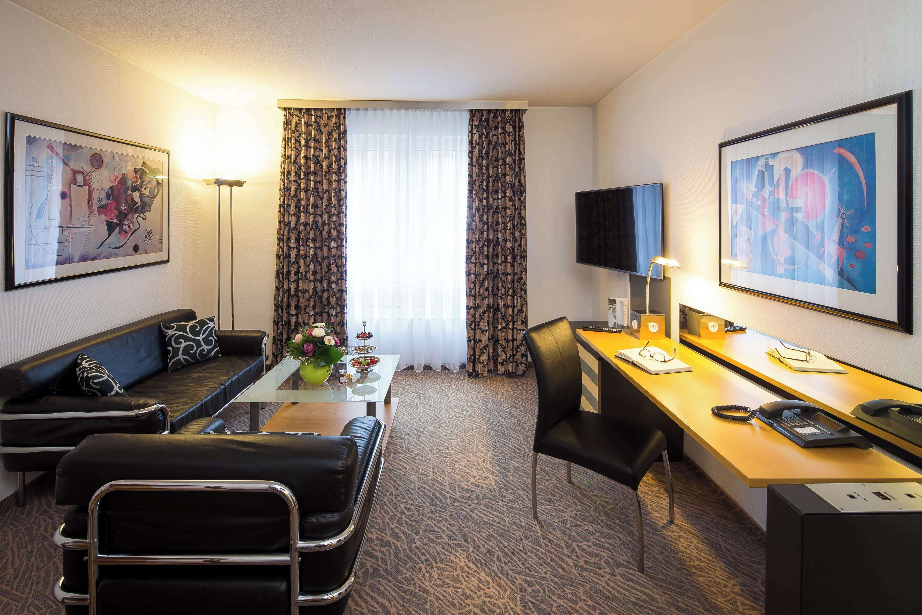 A hotel room with a sofa, a desk, a chair, a flat-screen TV, and artwork on the walls.
