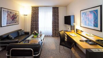 A hotel room with a sofa, a desk, a chair, a flat-screen TV, and artwork on the walls.