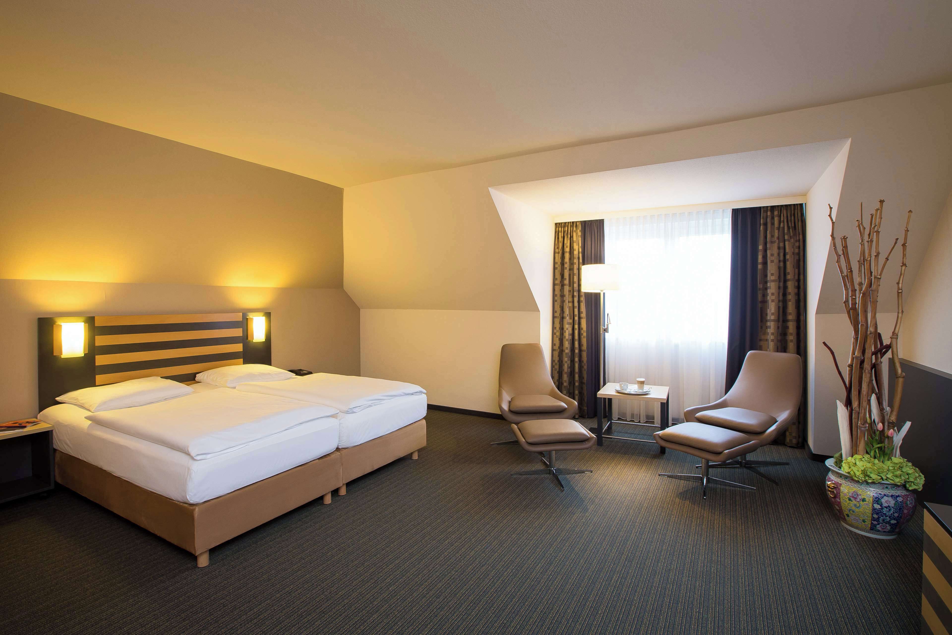 A hotel room with a double bed, two armchairs, a small table, and a potted plant.