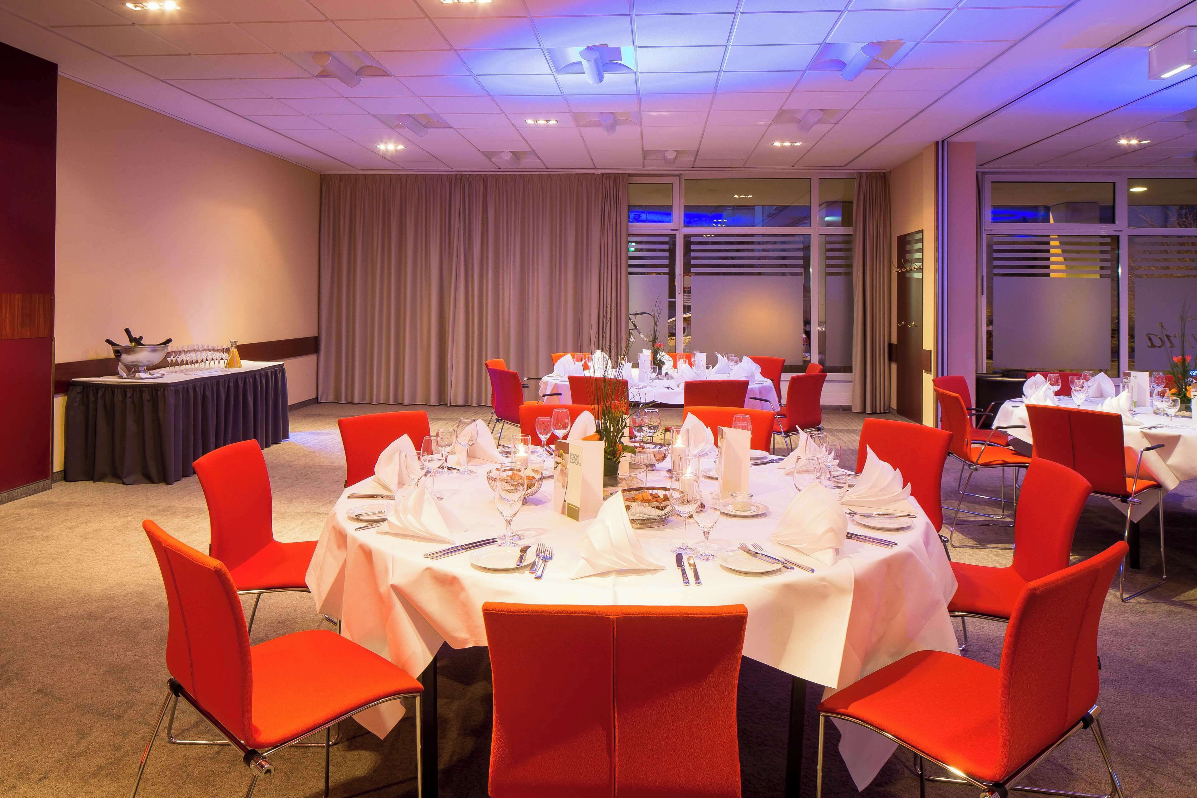 Un salón de conferencias con mesas redondas dispuestas para un evento formal, equipado con manteles blancos, sillas rojas y una variedad de vajilla.