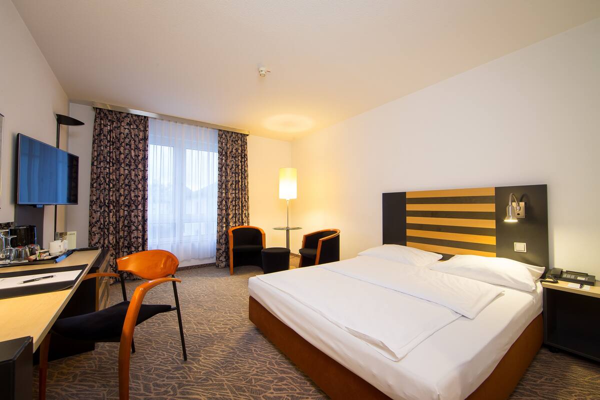 A hotel room with a bed, desk, chairs, a TV, and a lamp.