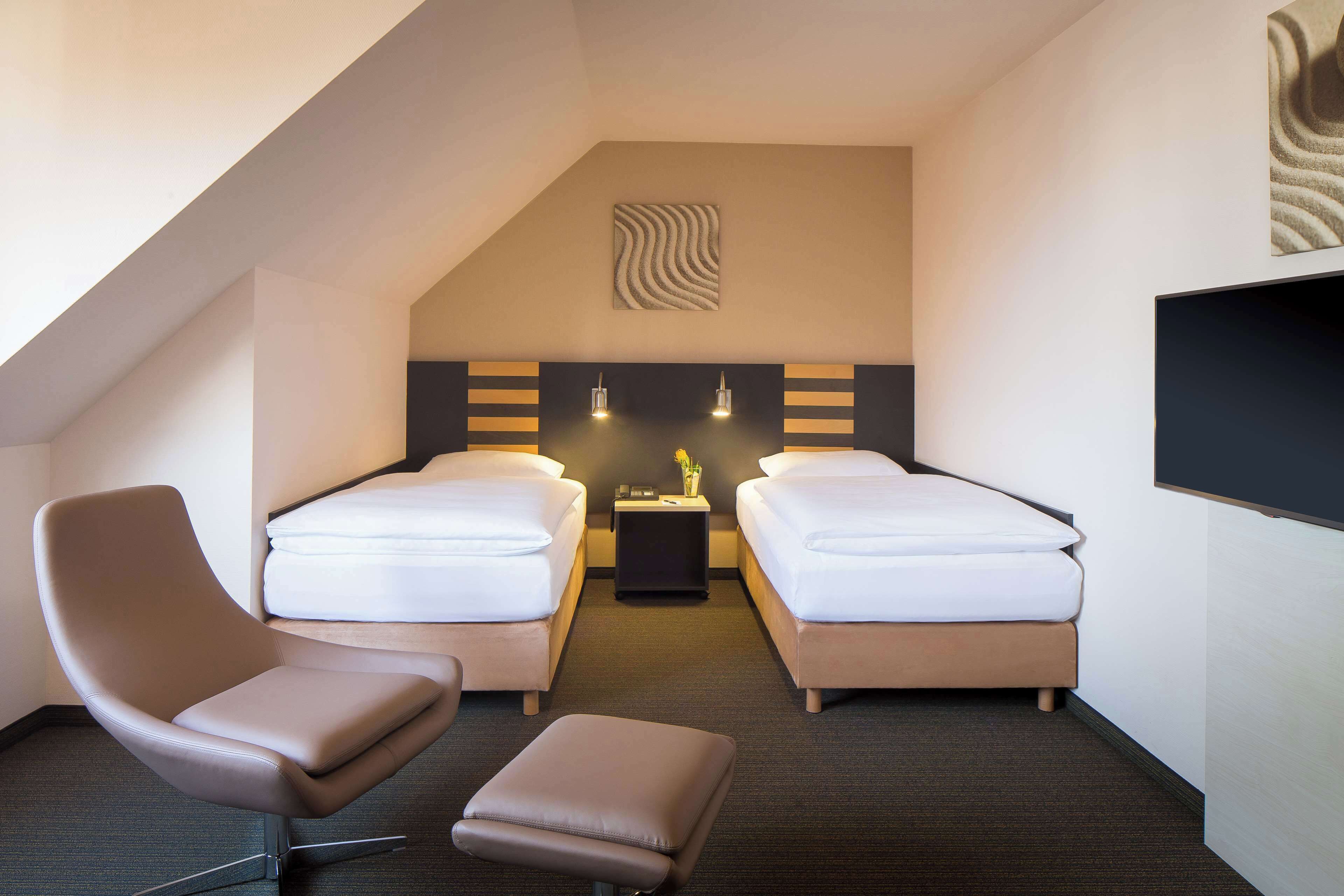 Ein Hotelzimmer mit zwei Betten, einem Sessel, einem Hocker, einem Nachttisch und einem an der Wand befestigten Fernseher.