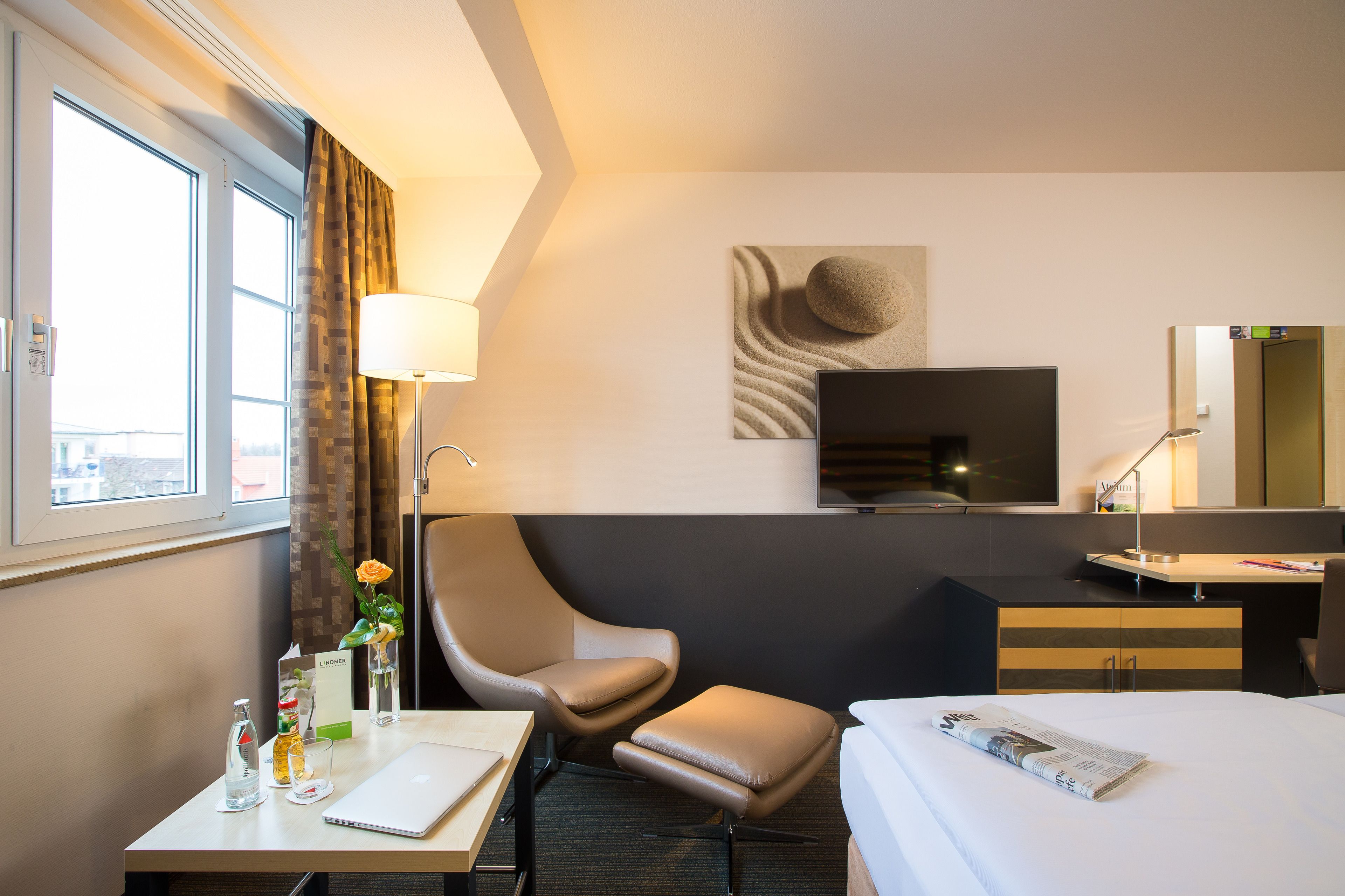 Ein Hotelzimmer mit Bett, Schreibtisch mit Laptop, Stuhl, Fernseher und einem Fenster mit Vorhängen.