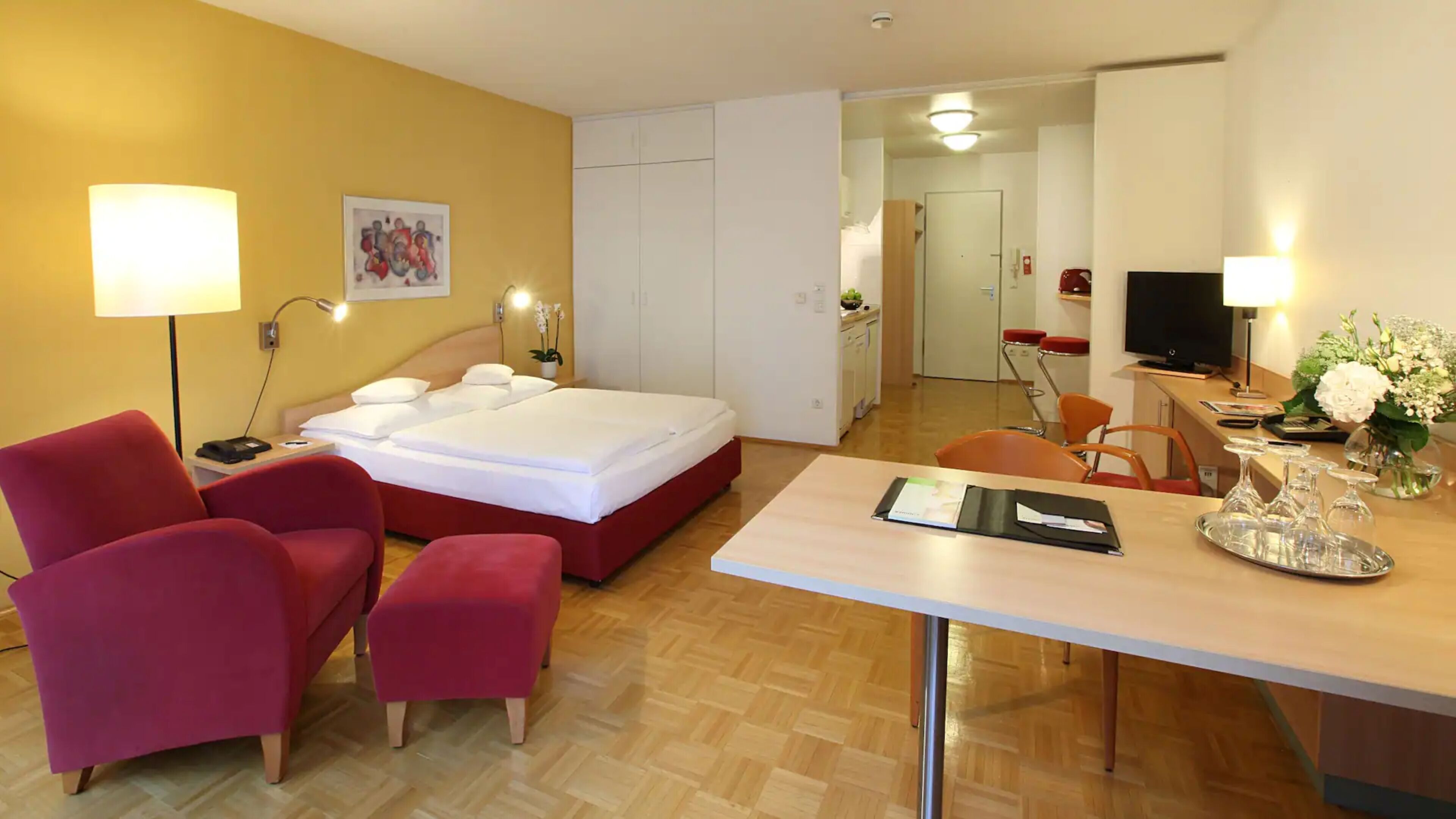 Quarto de hotel com cama, escrivaninha, cadeira, mesa pequena, abajur, televisão e um quadro na parede.