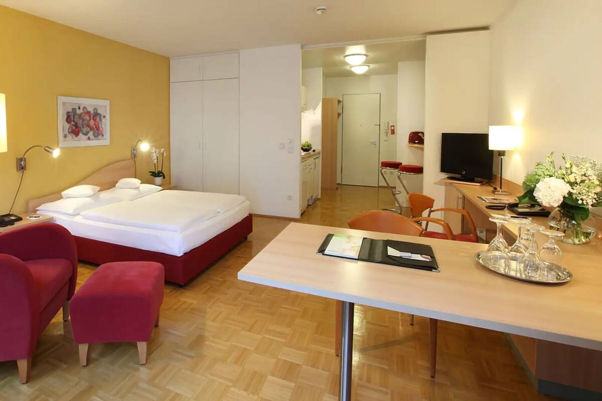 A hotel room with a bed, a desk, a chair, a small table, a lamp, a TV, and a painting on the wall.