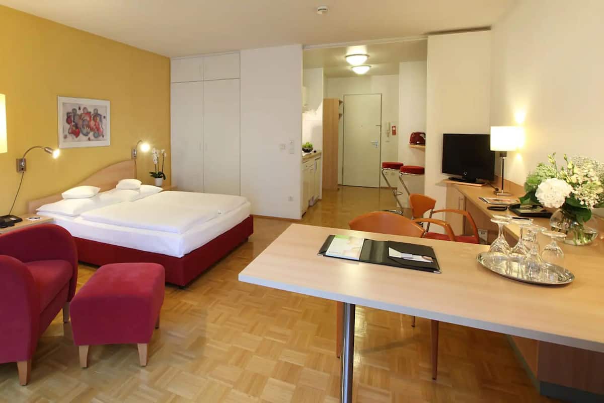 Habitación de hotel con cama, escritorio, silla, mesita, lámpara, televisor y un cuadro en la pared.