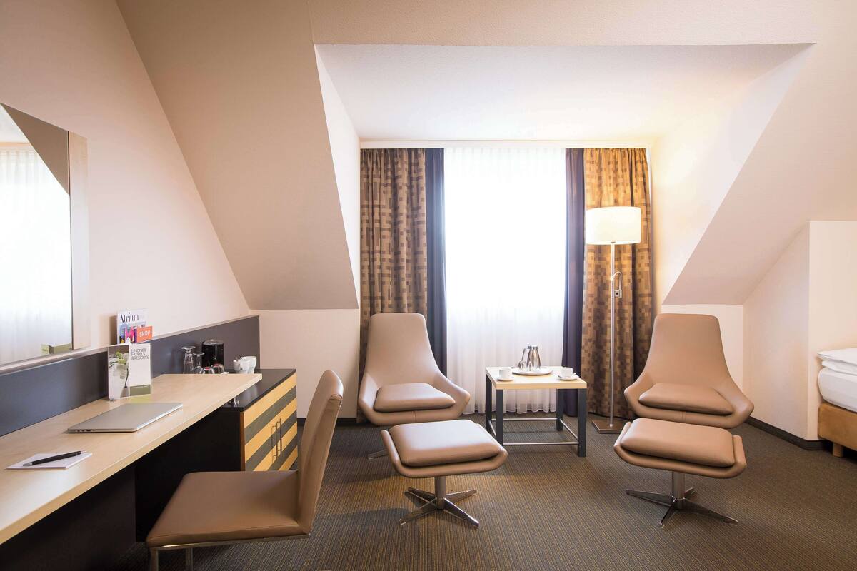 A hotel room with a desk, two armchairs, a bed, a small table with a lamp, and a window with curtains.