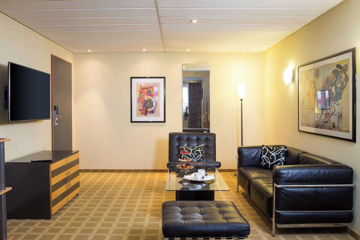 A modern living room with a flat-screen TV, a glass coffee table, a black leather sofa, and a black leather ottoman.