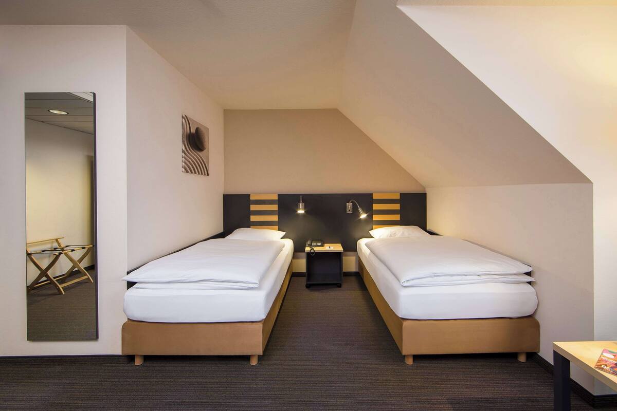 A hotel room with two single beds, a small table, a mirror, and a nightstand with a lamp.