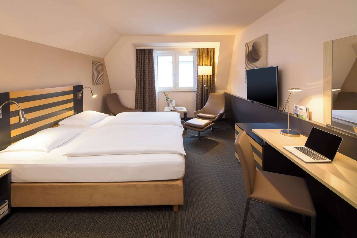 A hotel room with a large bed, a desk with a laptop, a TV, and a chair.