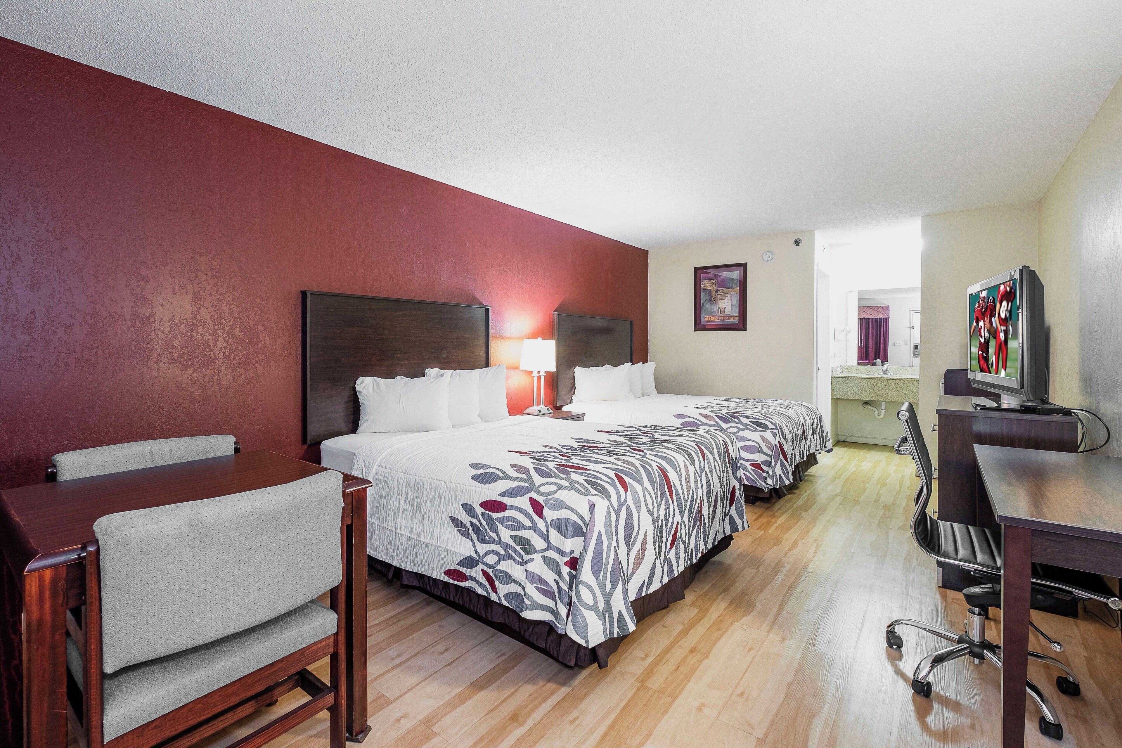 A hotel room with a bed, a desk, a chair, a TV, and a bathroom visible through an open door.