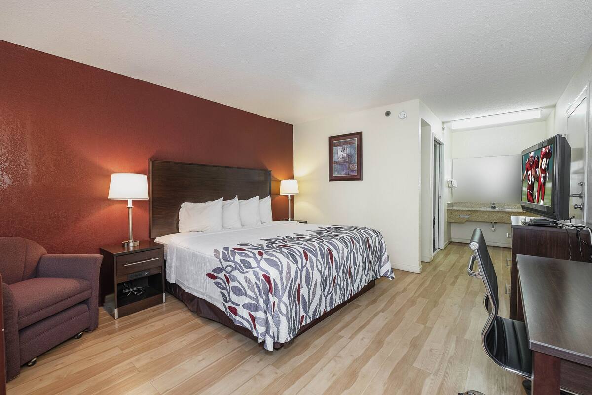 Quarto de hotel com cama, mesas de cabeceira, uma cadeira, escrivaninha e televisão.