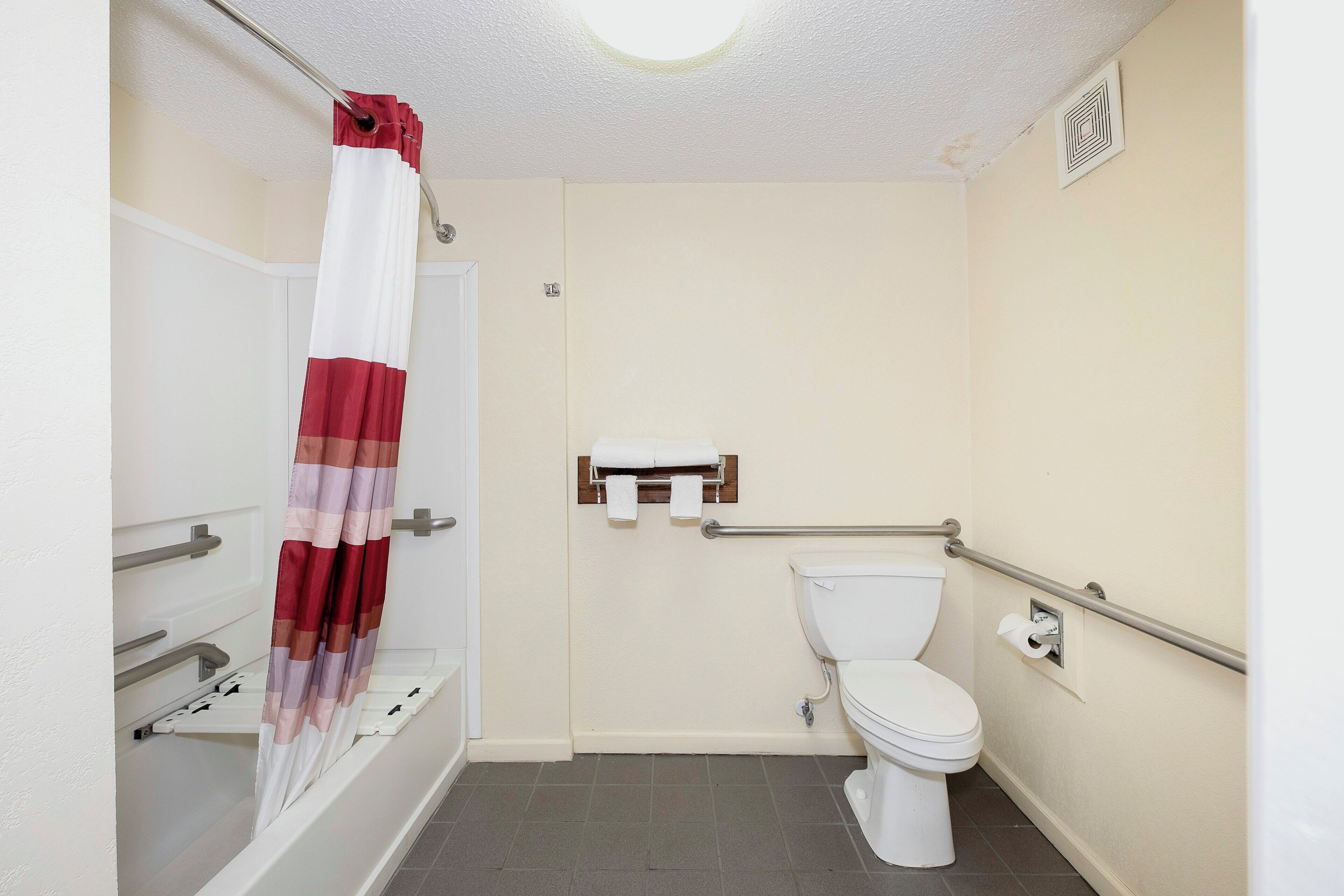 A bathroom with a white toilet, a shower with a red and white curtain, and a grab bar near the toilet.