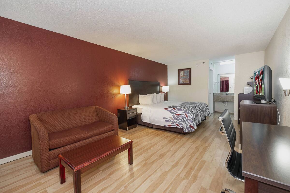 Quarto de hotel com cama, sofá, escrivaninha, televisão e banheiro ao fundo.