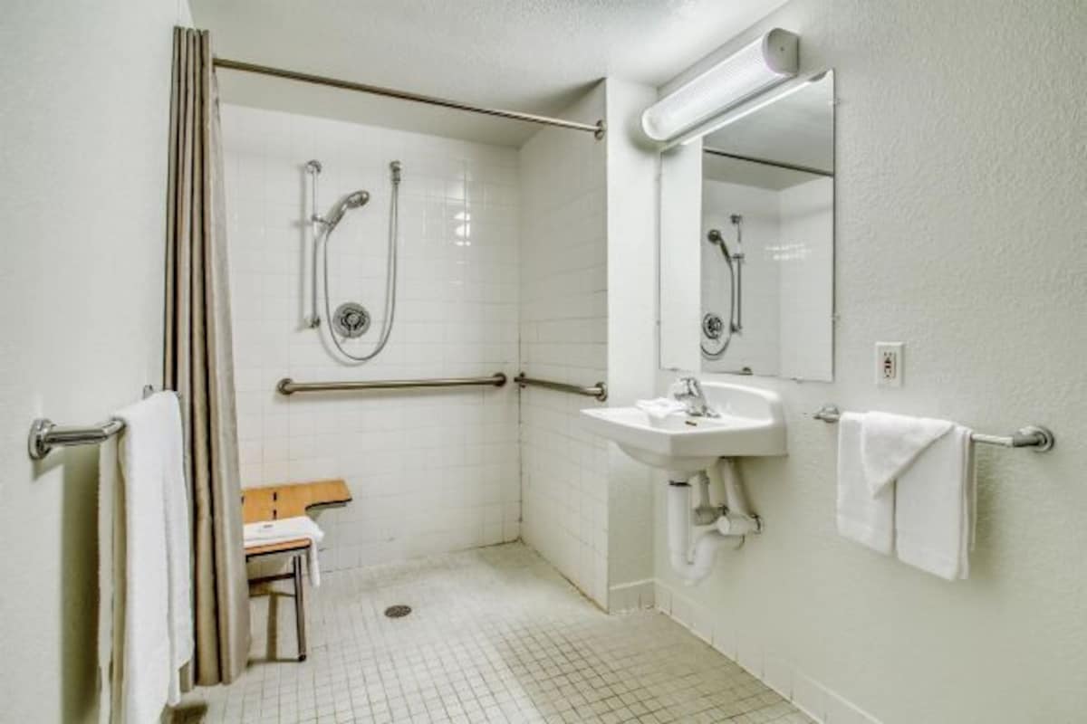 A small, white, wheelchair-accessible bathroom with a sink, mirror, and shower.