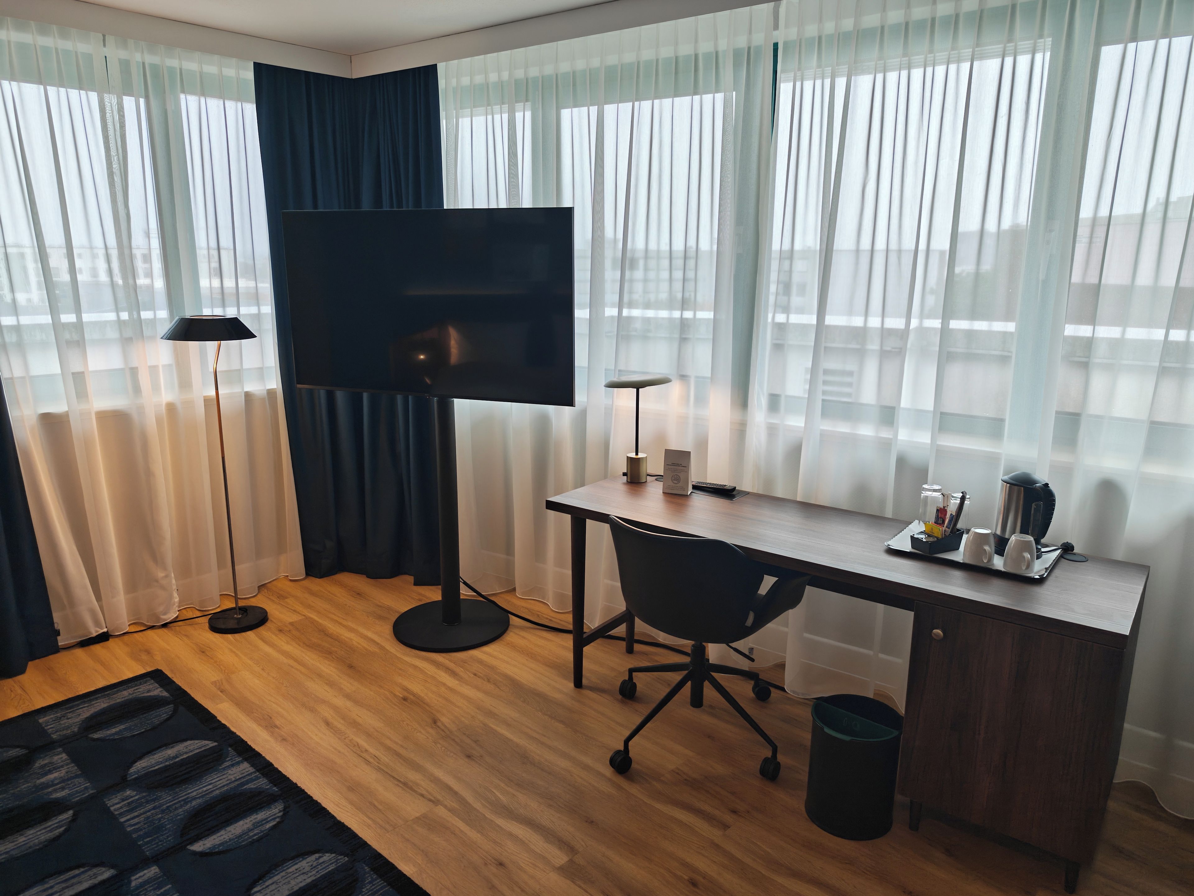 A modern hotel room with a flat-screen TV, a desk with a chair, a lamp, and a coffee station.