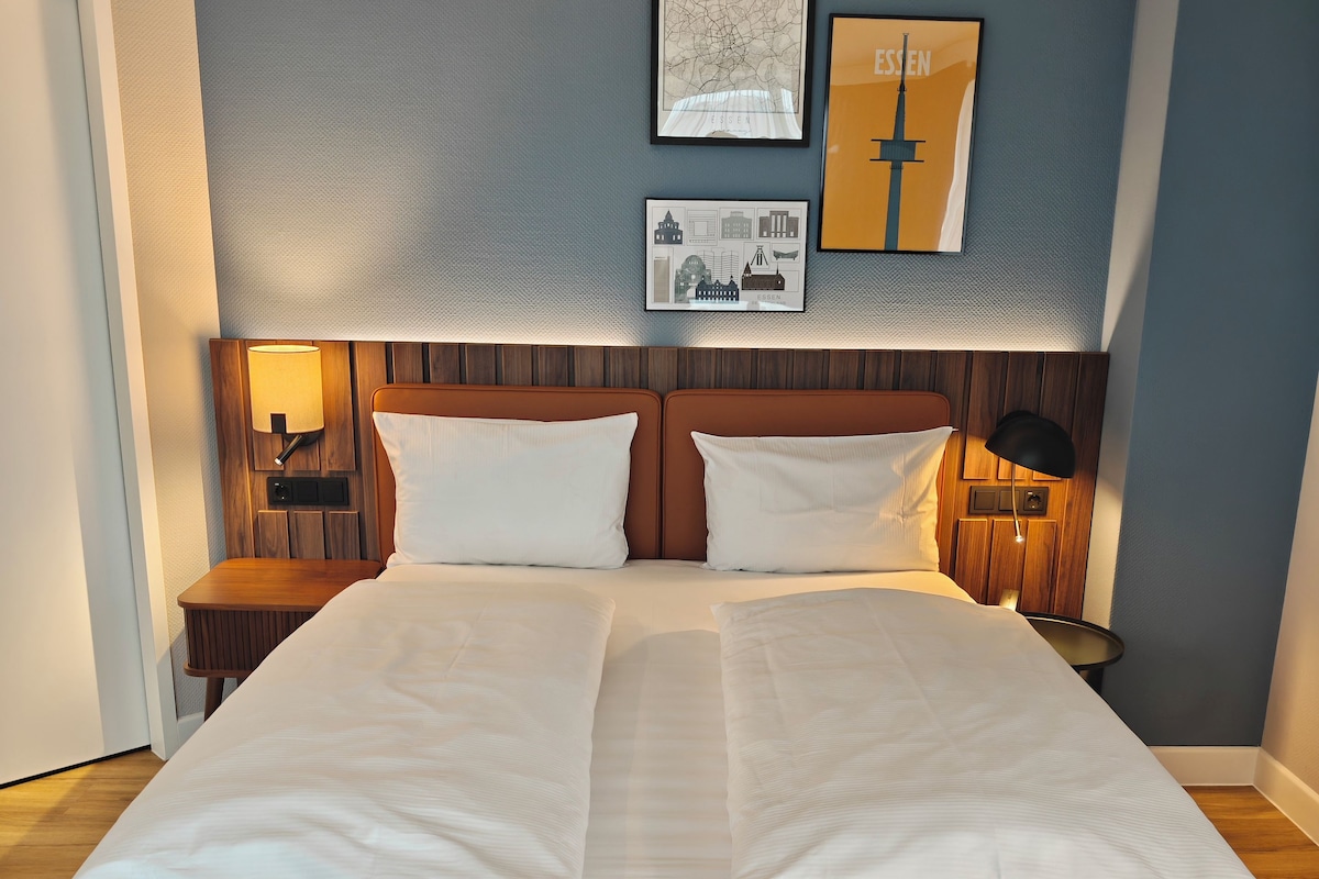Un lit double avec du linge de lit blanc, une tête de lit en bois et une table de chevet avec une lampe.