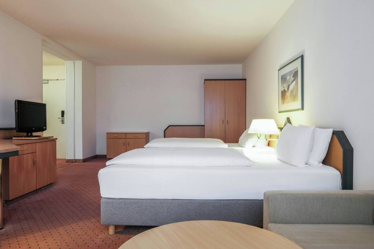 A hotel room with a bed, a desk with a television, a nightstand, a lamp, and a painting on the wall.