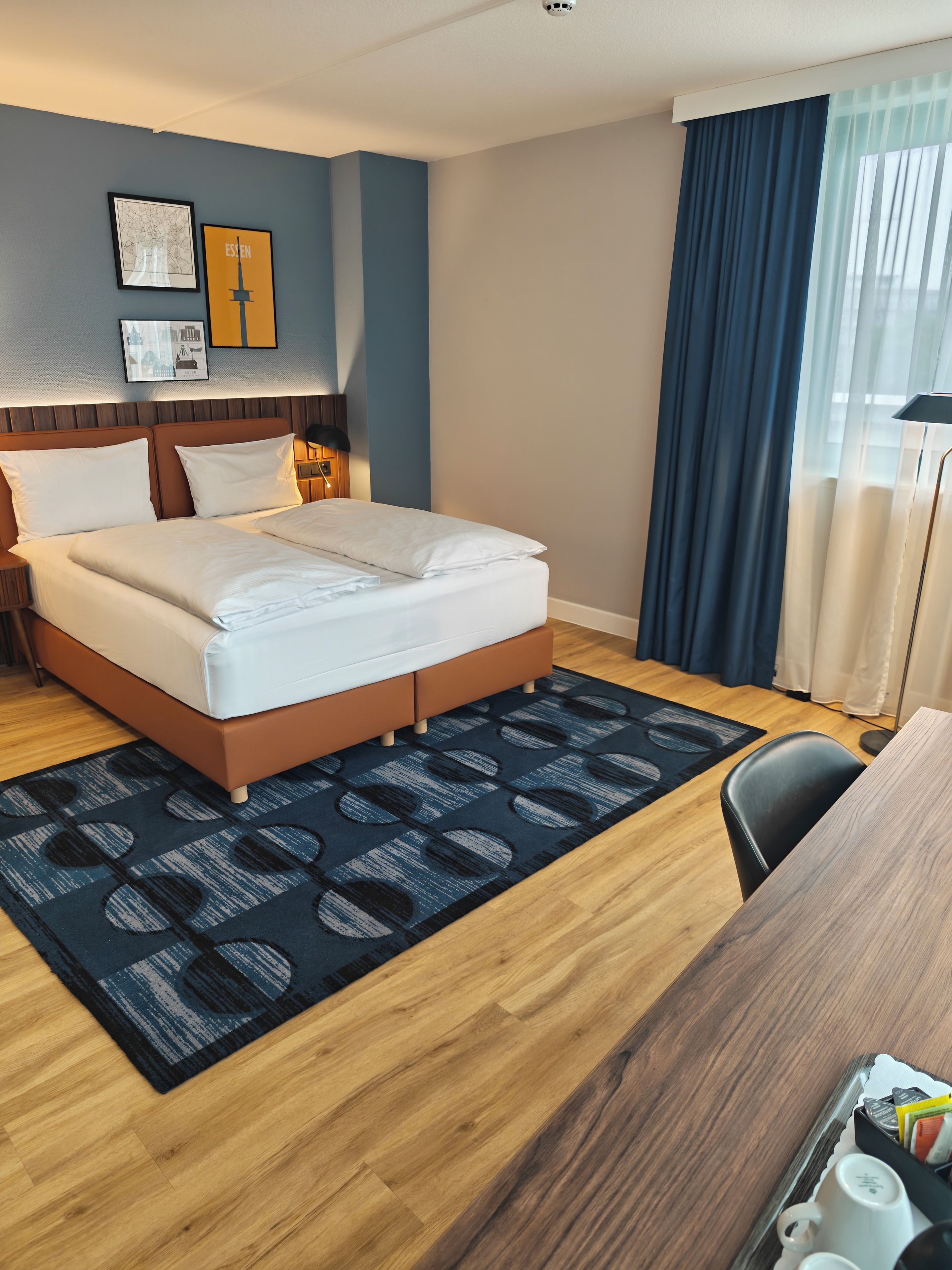 A hotel room with a bed, a desk, a chair, a lamp, and a window with curtains.