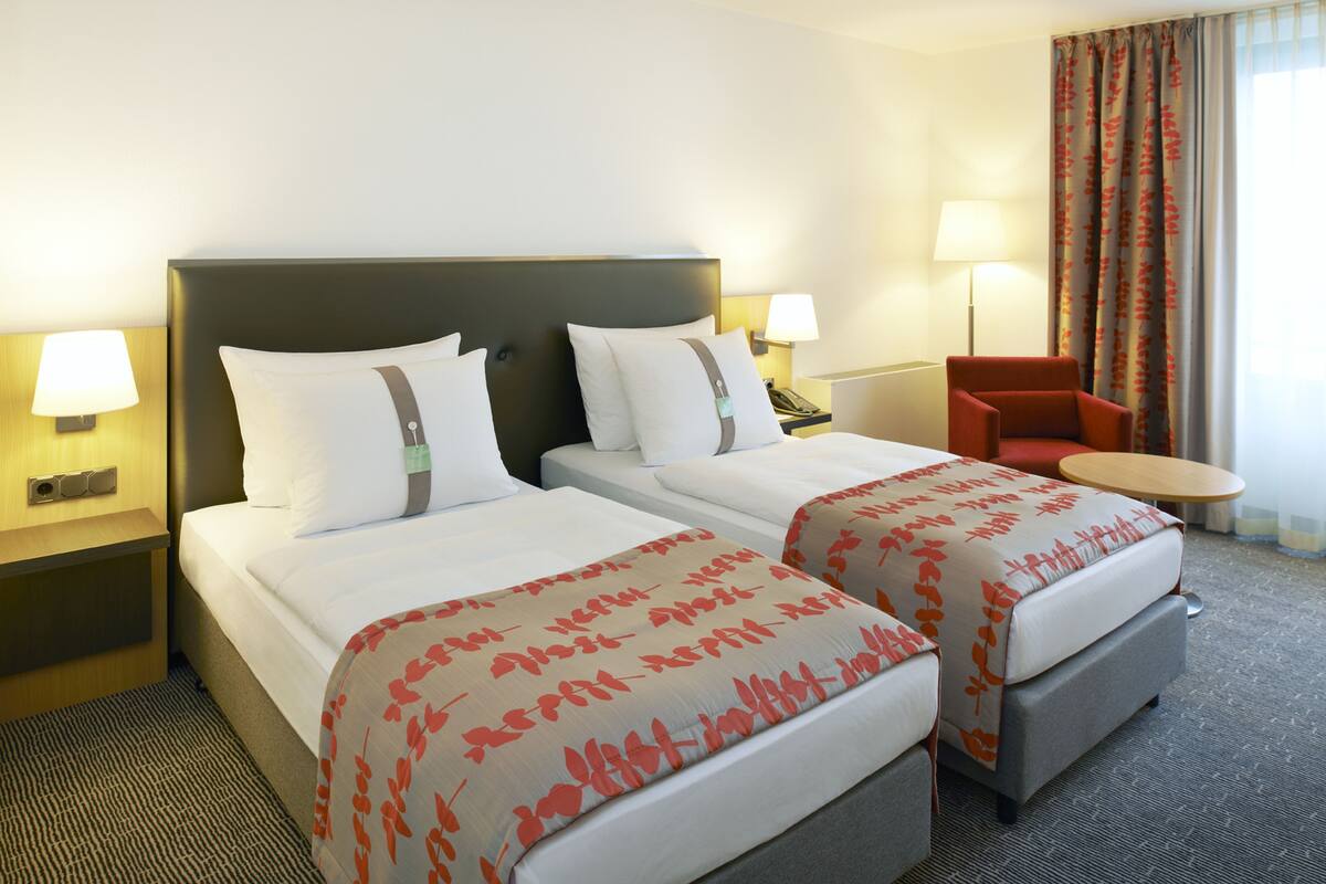 A hotel room with two beds, a small table, a chair, and a lamp.