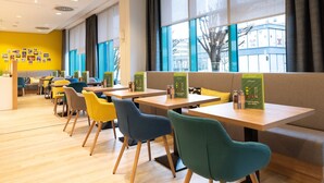 Un café moderne avec des tables en bois et des chaises colorées, de grandes fenêtres avec des rideaux, et un mur jaune orné de tableaux.