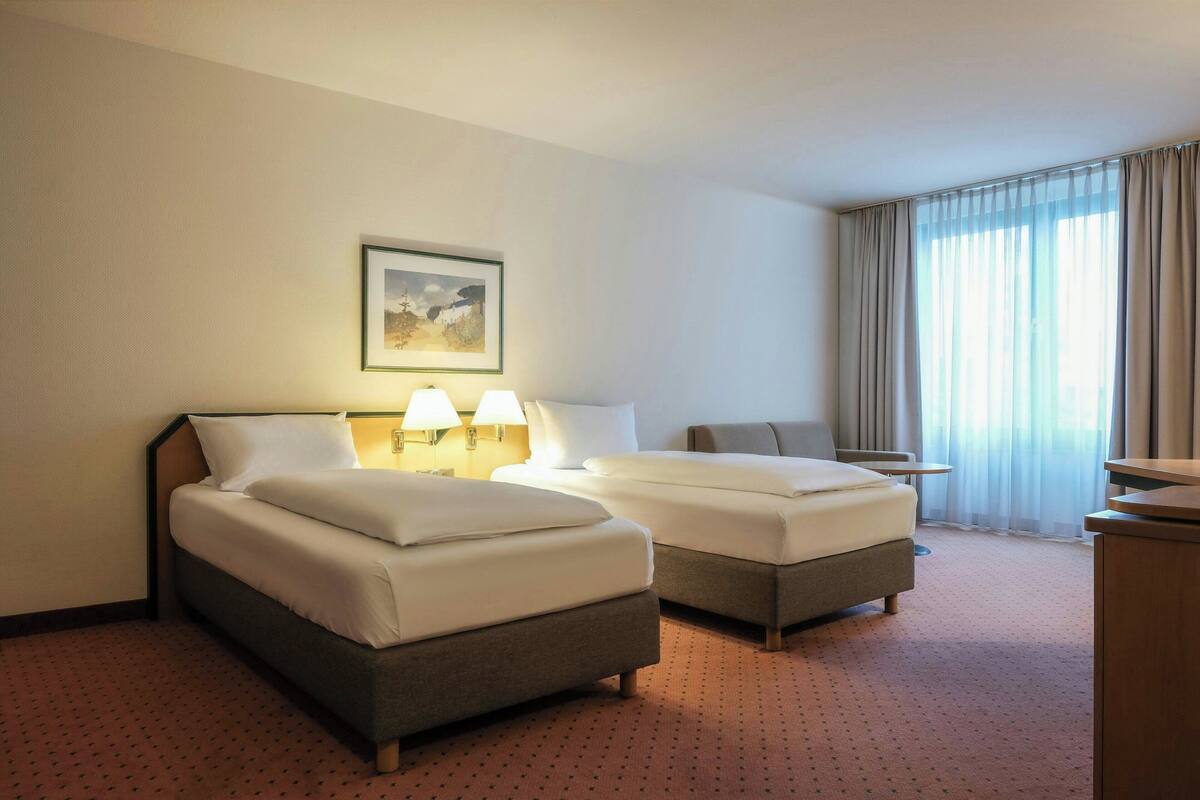 A hotel room with two beds, a desk, a chair, and a large window with curtains.