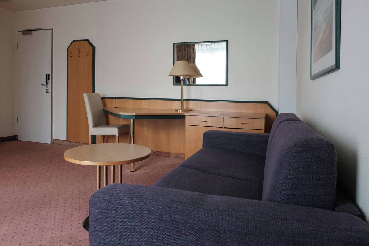 A room with a sofa, a small round table, a chair, a desk with a lamp, a wardrobe, and a door.