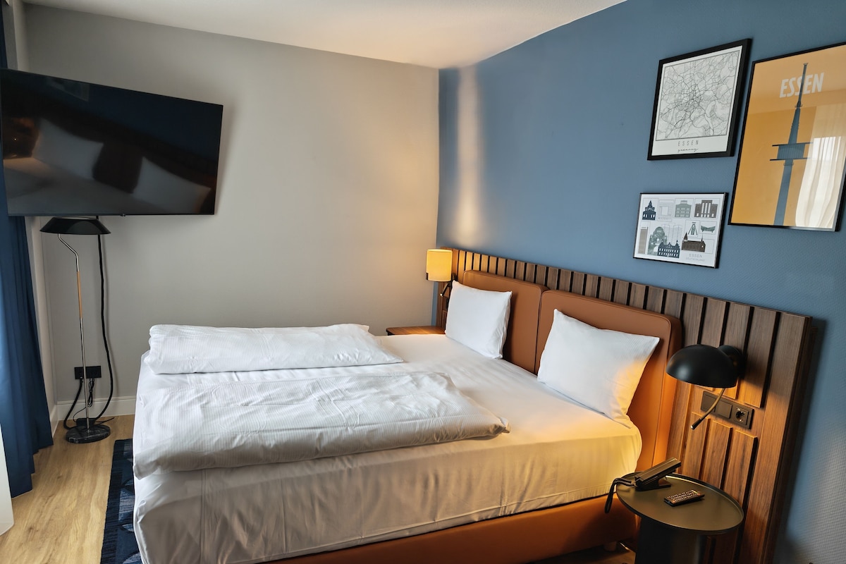 Une chambre d’hôtel avec un lit, une table de chevet avec une lampe, un téléviseur à écran plat fixé au mur et un tableau représentant une vue panoramique de la ville.