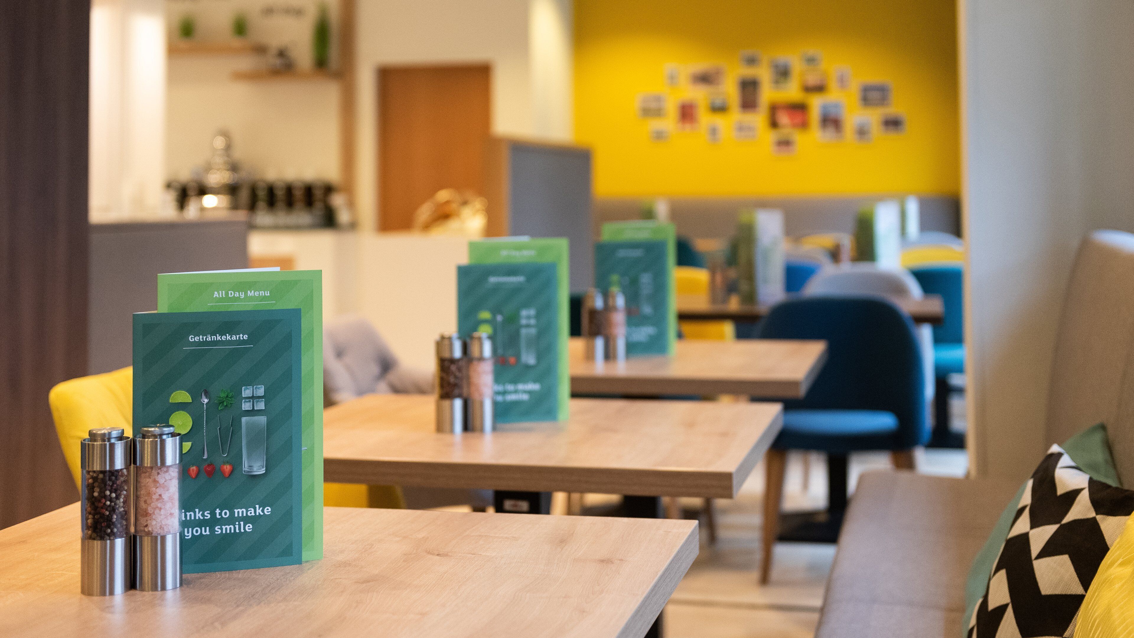 Un café moderne avec des tables et des chaises en bois, des tableaux de menu verts et un mur jaune orné de tableaux.
