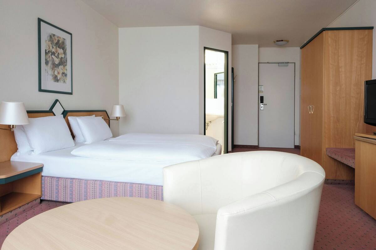 A hotel room with a bed, bedside tables, a chair, a round table, a television, and a door leading to another room.